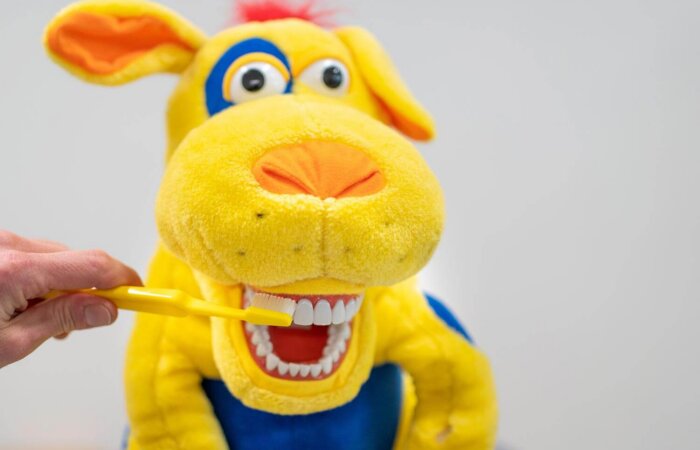 Gelbes Plüschtier mit Zähnen, dem eine Hand mit einer Zahnbürste die Zähne putzt Gelbes Plüschtier mit Zähnen, dem eine Hand mit einer Zahnbürste die Zähne putzt